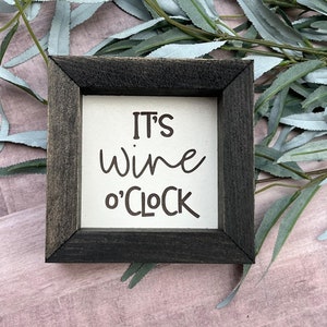 May include: A small wooden sign with a black frame and the text "It's wine o'clock" in brown lettering.