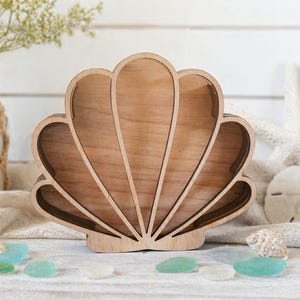 May include: Wooden seashell-shaped tray with a natural finish. The tray is surrounded by sea glass, seashells, and a starfish. A vase with flowers is in the background. The tray is designed to hold small items.