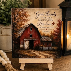 Pode incluir: Uma placa quadrada de madeira com uma ilustração impressa de um celeiro vermelho rodeado por árvores de outono. O texto diz: Give thanks TO THE LORD, FOR HE IS GOOD; His love ENDURES FOREVER. PSALM 107:1. A placa está num pequeno cavalete.