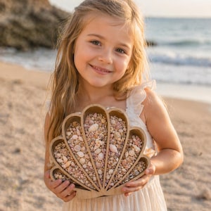 May include: A wooden seashell-shaped tray filled with small shells and pebbles. The tray is held by a person on a sandy beach. The tray is made of light-colored wood and has a detailed, scalloped edge.