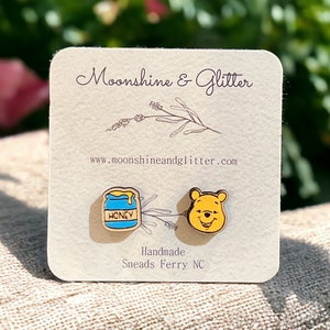 May include: A pair of wooden earrings featuring a Winnie the Pooh character and a honey pot. The earrings are on a card that says "Moonshine & Glitter" and "Handmade, Sneads Ferry NC".