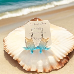 May include: A pair of blue and yellow sea turtle earrings with silver hooks. The earrings are on a white card with the text "Moonshine & Glitter" and a website address.