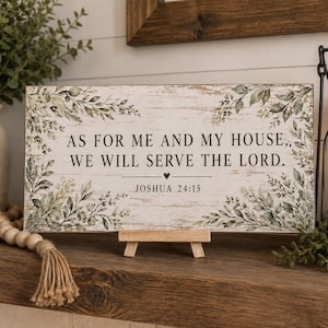 Könnte beinhalten: Ein rechteckiges Holzschild mit weißer Used-Optik und grünen botanischen Illustrationen in den Ecken. Der Text lautet: AS FOR ME AND MY HOUSE, WE WILL SERVE THE LORD. JOSHUA 24:15, mit einem kleinen Herzsymbol unter dem Text zentriert.