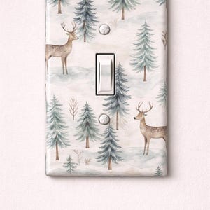 Pode incluir: Uma capa decorativa para interruptor de luz com um design de floresta de inverno. O design inclui ilustrações em aquarela de veados, árvores perenes e uma paisagem nevada em tons de verde, castanho e branco. O interruptor é branco.