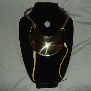 May include: A crescent-shaped polished brass gorget necklace featuring a strand of dark purple beads with gold-tone spacers. The piece includes two light tan leather cords hanging from the sides and is displayed with a silver-colored coin.