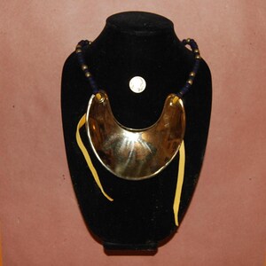 May include: A gold-toned, crescent-shaped necklace with a dark blue beaded strand and yellow leather ties. The necklace is displayed on a black bust against a brown background. A silver coin is also visible.