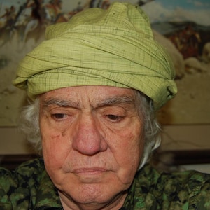 May include: A light green head covering featuring a subtle grid pattern and multiple horizontal folds wrapped around the crown. The fabric has a textured appearance with visible weave lines throughout the material, creating a layered look.