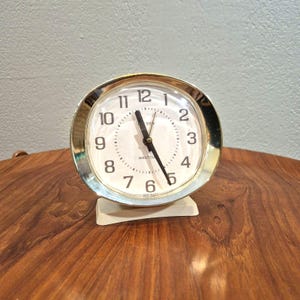 May include: A vintage Westclox Ben alarm clock with a cream-colored face and gold-tone metal frame. The clock has black numerals and hands, and sits on a cream-colored base. The clock is on a wooden surface.