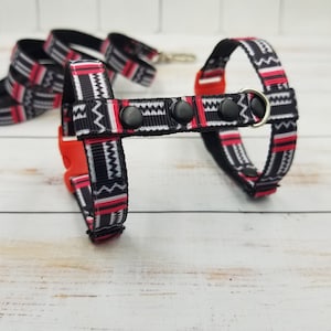 Può includere: Un'imbracatura per cani con motivo nero, bianco e rosso con un anello a D argentato e fibbie in plastica nera.