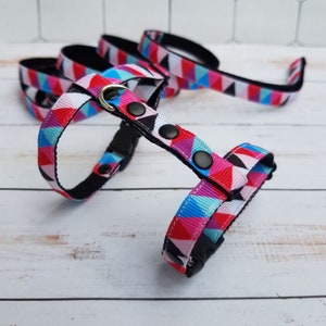 May include: A colorful geometric pattern dog harness with black webbing and a silver D-ring. The harness has adjustable straps and a buckle closure.