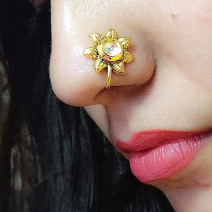 Puede incluir: Un aro para la nariz de color dorado con un diseño de flor y una piedra transparente en el centro.