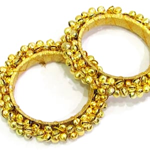 Può includere: Due braccialetti dorati con piccoli campanellini dorati attorno alla circonferenza. I braccialetti sono in tessuto e sono di colore oro chiaro.