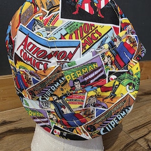 May include: A fabric cap featuring a collage of vintage comic book covers. The design includes panels with the words "Action Comics" and "Superman," in vibrant colors. The cap is displayed on a white form.