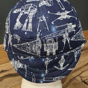 May include: A navy blue welding cap featuring a white schematic design of spaceships and vehicles against a starry background. The cap is made of fabric and is designed to protect the wearer's head.
