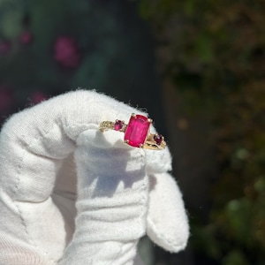 May include: A gold ring featuring a large, rectangular, bright pink gemstone as the centerpiece, flanked by smaller, round, red gemstones. The ring has a delicate, ornate design with additional small, round gemstones.