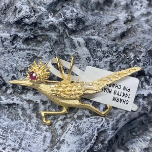 May include: A gold-colored roadrunner charm with a red gemstone eye. The charm is attached to a white tag that reads "CHARM 1AKTYG ROAD PIN CHAP".