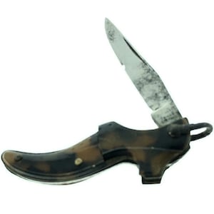 May include: A small, novelty pocket knife shaped like a high-heeled shoe. The shoe is a mix of brown and black, with a silver blade extended upwards. The blade has a worn, textured appearance.