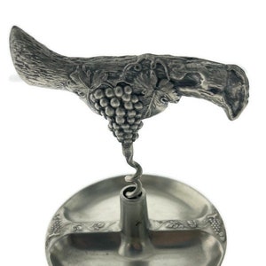 May include: A silver-tone metal corkscrew with a grapevine design. The corkscrew has a handle shaped like a grapevine with a bunch of grapes. The corkscrew is attached to a round base with a small dish.