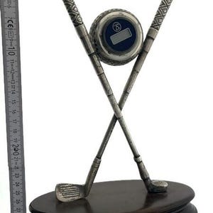 May include: A silver-toned metal golf trophy featuring two crossed golf clubs and a golf ball with a digital clock face, mounted on an oval wooden base. A vertical ruler next to the object shows a height of approximately 33 centimetres.