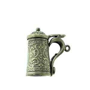 Puede incluir: Un amuleto de metal plateado en forma de jarra de cerveza con un diseño ornamentado. El amuleto tiene una tapa con bisagras y un bucle para sujetarlo a una pulsera o collar.