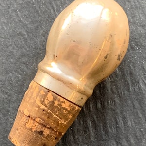 May include: A vintage bottle stopper with a pearlescent, bulbous top and a natural cork base. The metallic top has a slightly aged appearance. The cork base shows natural wood grain and texture. This decorative stopper is suitable for wine bottles.