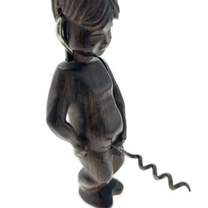 May include: A wooden figurine of a person holding a corkscrew in their hand. The figurine is dark brown and has a detailed face. The corkscrew is silver and has a spiral shape.