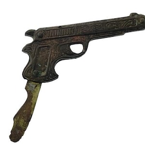 Puede incluir: Un abridor de botellas de estilo vintage con forma de pistola. El abridor está hecho de metal oscuro y envejecido con intrincados patrones florales y geométricos. El mango es de un color más claro y desgastado, lo que sugiere edad y uso.