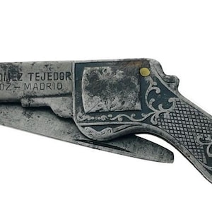 Puede incluir: Una navaja plegable vintage con forma de pistola, con acabado plateado y detalles decorativos intrincados. La hoja está parcialmente extendida. El texto "JOSE GOMEZ TEJEDOR BADAJOZ - MADRID" está grabado en el mango.