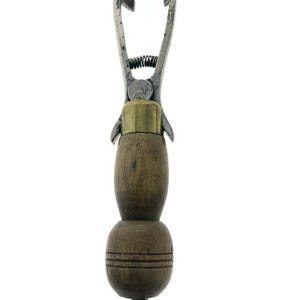 May include: Antique metal and wood tool with a spring-loaded clamp and a wooden handle. The handle is made of two sections, one round and one oval, with a brass ring at the top.