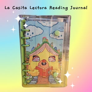 May include: A clear plastic journal with a green spine and button closures. The cover features the text "La Casita Lectora Reading Journal" and a colorful illustration of a house reading a book. The journal is designed for reading.