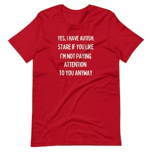 Puede incluir: Una camiseta roja con texto blanco. El texto dice: "YES, I HAVE AUTISM. STARE IF YOU LIKE I'M NOT PAYING ATTENTION TO YOU ANYWAY." La camiseta tiene cuello redondo y mangas cortas.