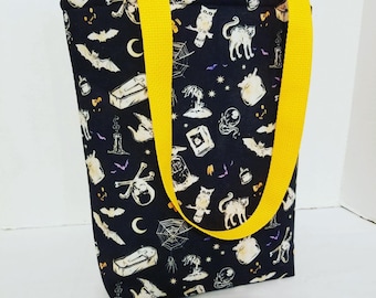 May include: A black tote bag with a white and yellow Halloween-themed print. The bag has yellow straps and features images of bats, owls, cats, skulls, and other spooky elements.