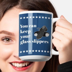 May include: White ceramic mug with a blue background and white text that reads "You can keep your glass slippers". The mug has a black and white illustration of a pair of tap shoes.