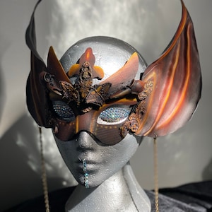 May include: A masquerade mask with intricate details, featuring a brown and orange colour scheme with a gold chain. The mask has a winged design and is adorned with rhinestones.