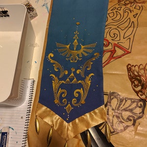 K&ouml;nnte beinhalten: Eine teal- und goldfarbene Zierfahne mit einem goldgestickten Design eines Vogels und einer Krone. Die Fahne hat einen goldenen Satinrand und goldene, tropfenf&ouml;rmige Verzierungen. Handgefertigt.