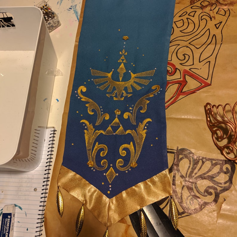 K&ouml;nnte beinhalten: Eine teal- und goldfarbene Zierfahne mit einem goldgestickten Design eines Vogels und einer Krone. Die Fahne hat einen goldenen Satinrand und goldene, tropfenf&ouml;rmige Verzierungen. Handgefertigt.