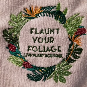 K&ouml;nnte beinhalten: Wei&szlig;es Handtuch mit gesticktem Text "FLAUNT YOUR FOLIAGE", umgeben von einem Kranz aus gr&uuml;nen, gelben und roten tropischen Bl&auml;ttern und Blumen. Der Text "LIVE PLANT BOUTIQUE" befindet sich darunter.