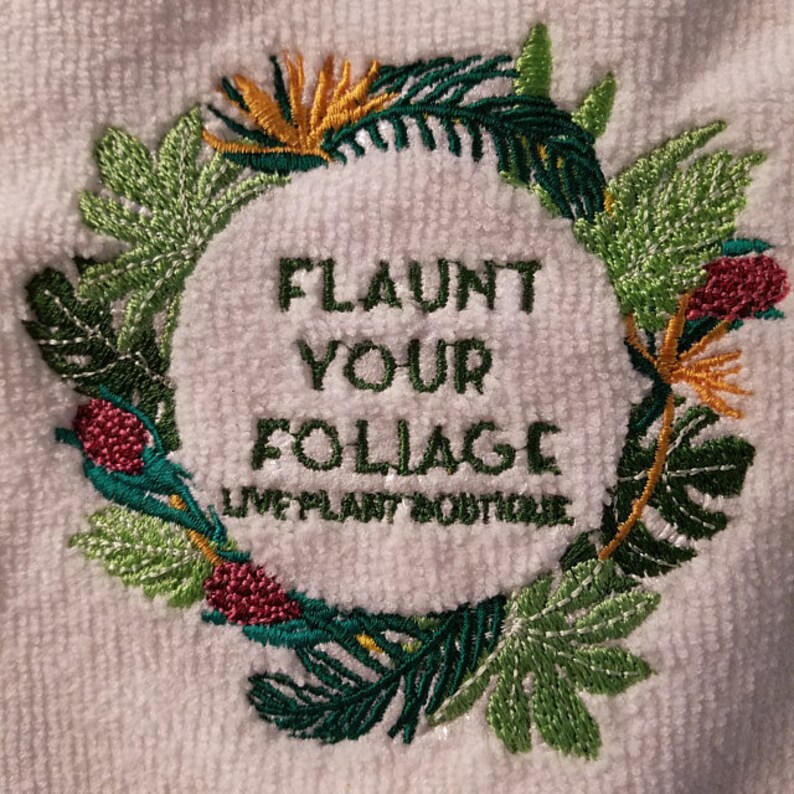 K&ouml;nnte beinhalten: Wei&szlig;es Handtuch mit gesticktem Text "FLAUNT YOUR FOLIAGE", umgeben von einem Kranz aus gr&uuml;nen, gelben und roten tropischen Bl&auml;ttern und Blumen. Der Text "LIVE PLANT BOUTIQUE" befindet sich darunter.