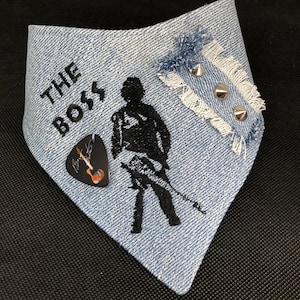 May include: A blue denim bandana with the text "THE BOSS" embroidered on it. A black silhouette of a person playing a guitar is also embroidered on the bandana. A guitar pick is attached to the bandana. The bandana has a frayed edge and a small piece of denim with silver studs is attached to the top.