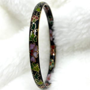 May include: A thin, circular bangle bracelet featuring a black enamel base with gold-toned metal wire outlines. The surface is decorated with a repeating floral pattern in shades of light purple, dark green, and red, creating a continuous design.