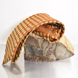 May include: A necktie in a rich burnt orange hue featuring a repeating geometric pattern of small, intricate yellow and white floral-like motifs. The fabric has a subtle sheen, and the tie is draped over a textured, multi-colored stone surface.