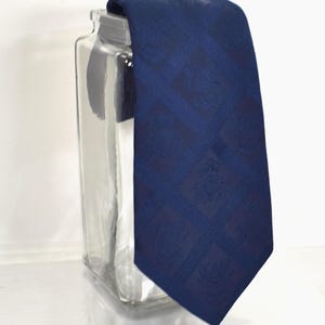 May include: A dark blue necktie featuring a repeating diamond pattern with subtle crest-like emblems centered within each diamond. The fabric has a woven texture and a slight sheen, draped over a clear glass container with a rectangular shape.