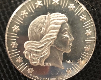 May include: A silver coin featuring a profile of a woman with flowing hair and a laurel wreath. The coin's edge is decorated with stars and lines. The text "WORLD WIDE MINT, INC. 1981" is inscribed along the bottom edge.