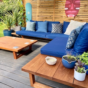 Puede incluir: Juego de muebles de jardín de madera con cojines azules. Incluye una mesa de centro cuadrada y un sofá seccional. Cojines decorativos y plantas en macetas añaden color. Una pantalla de madera y una tabla de surf están en el fondo. La mesa de centro mide unos 90 cm x 90 cm.