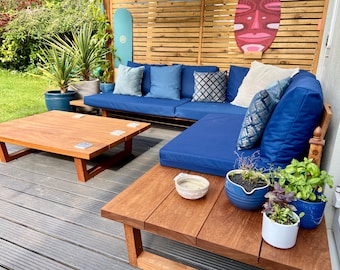 May include: Wooden outdoor furniture set with blue cushions. Features a square coffee table and a sectional sofa. Decorative cushions and potted plants add colour. A wooden screen and a surfboard are in the background. The coffee table is approximately 90cm x 90cm.