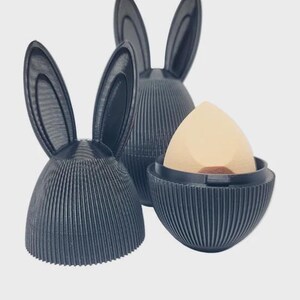 May include: Three black, ribbed, bunny-shaped holders. One holds a beige makeup sponge. The holders have tall, upright ears. The holders are made of a dark, matte material. The background is a plain, light color.