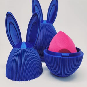 May include: Three blue bunny-shaped containers with vertical ribbed detailing. One container holds a pink makeup sponge. The bunny ears are upright. The containers are on a white background.
