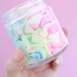 May include: A clear glass jar filled with pastel-colored, swirled whipped cream. The colors include pink, blue, green, and white. The jar is held against a soft pink background.