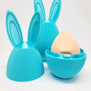 May include: Three turquoise bunny-shaped holders, one open with a beige makeup sponge inside. The holders have a ribbed texture and are designed with long, upright ears. The holders are designed to store and protect makeup sponges.