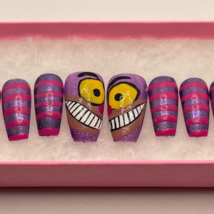 K&ouml;nnte beinhalten: Ein Set aus k&uuml;nstlichen Fingern&auml;geln mit einer lila Glitzerbasis und rosa Streifen sowie Designs der Grinsekatze. Die Grinsekatzen-N&auml;gel haben eine lila Basis, gelbe Augen und ein breites Grinsen. Die N&auml;gel werden in einer rosa Schachtel pr&auml;sentiert.